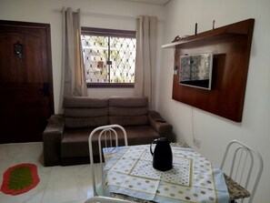 Un petit coin salon avec un canapé, une table sur laquelle se trouve une bouilloire, un téléviseur fixé au mur et une fenêtre avec des rideaux.