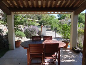 Un patio cubierto con una mesa redonda y sillas, rodeado de plantas en macetas y un muro de piedra.