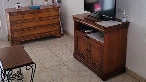 Una stanza con un comò in legno, una televisione su un mobile, un tavolino e una finestra con persiane.