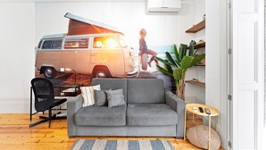Un salón con un sofá gris, una mesita redonda pequeña, una silla, un mural de una furgoneta y una planta.