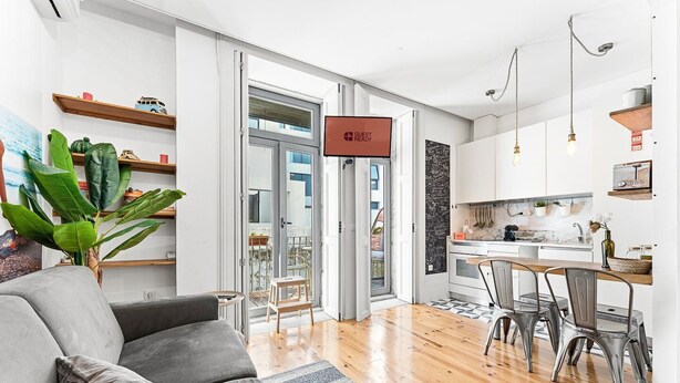 Una sala moderna con un sofá gris, una mesa de centro y una planta grande. Cuenta con una zona de cocina con una mesa de comedor y sillas, y un letrero que indica 'GUEST READY'.