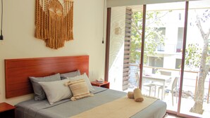 Cama bem arrumada com cabeceira de madeira, um painel de macramé na parede e vista para uma varanda externa com mesa e cadeiras.