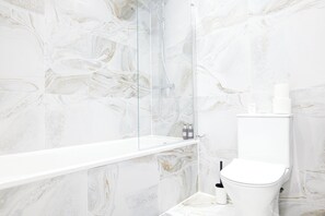 Ein Badezimmer mit Marmorwand, einer Glaskabine zur Dusche, einer weißen Badewanne und einer Toilette.