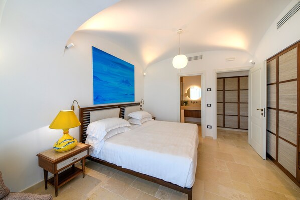 A bedroom with a bed, a nightstand, a lamp, a painting on the wall, and a wardrobe. - OraBlu Villas (Forio)