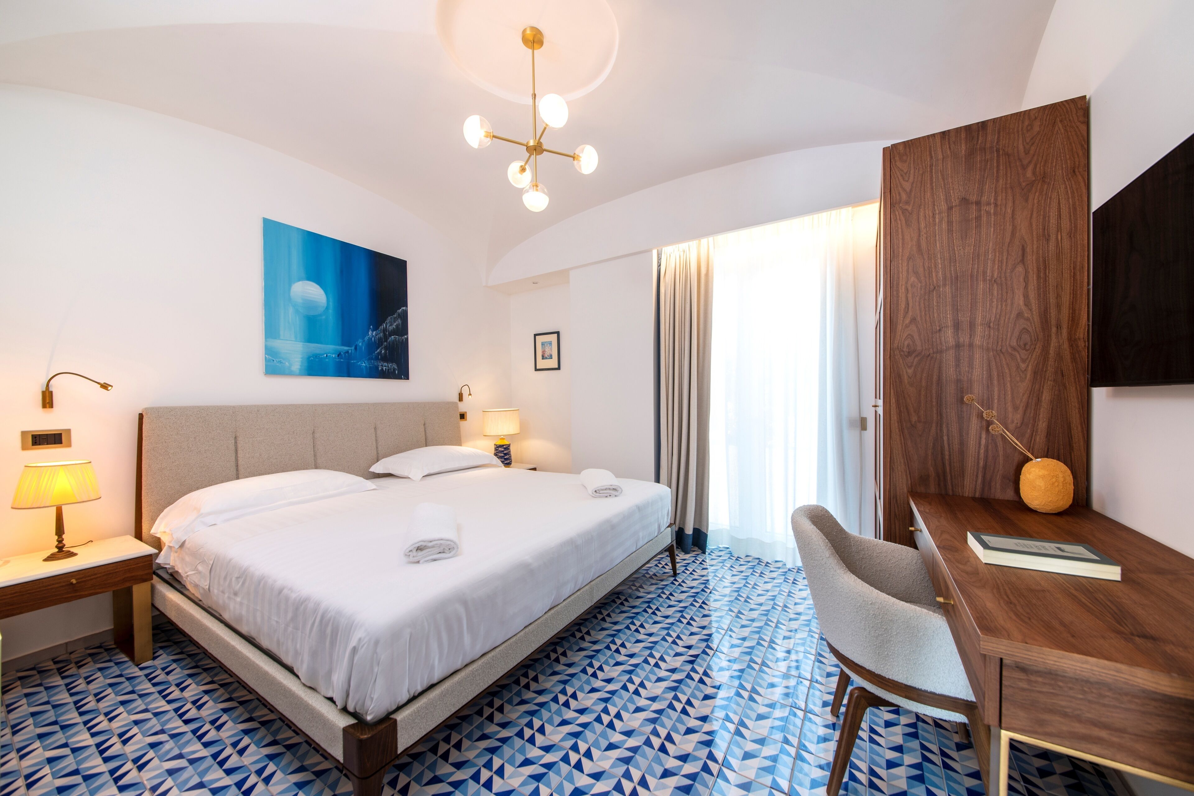 Habitación de hotel con una cama grande, un escritorio, una silla, una lámpara, un cuadro y vistas al exterior.