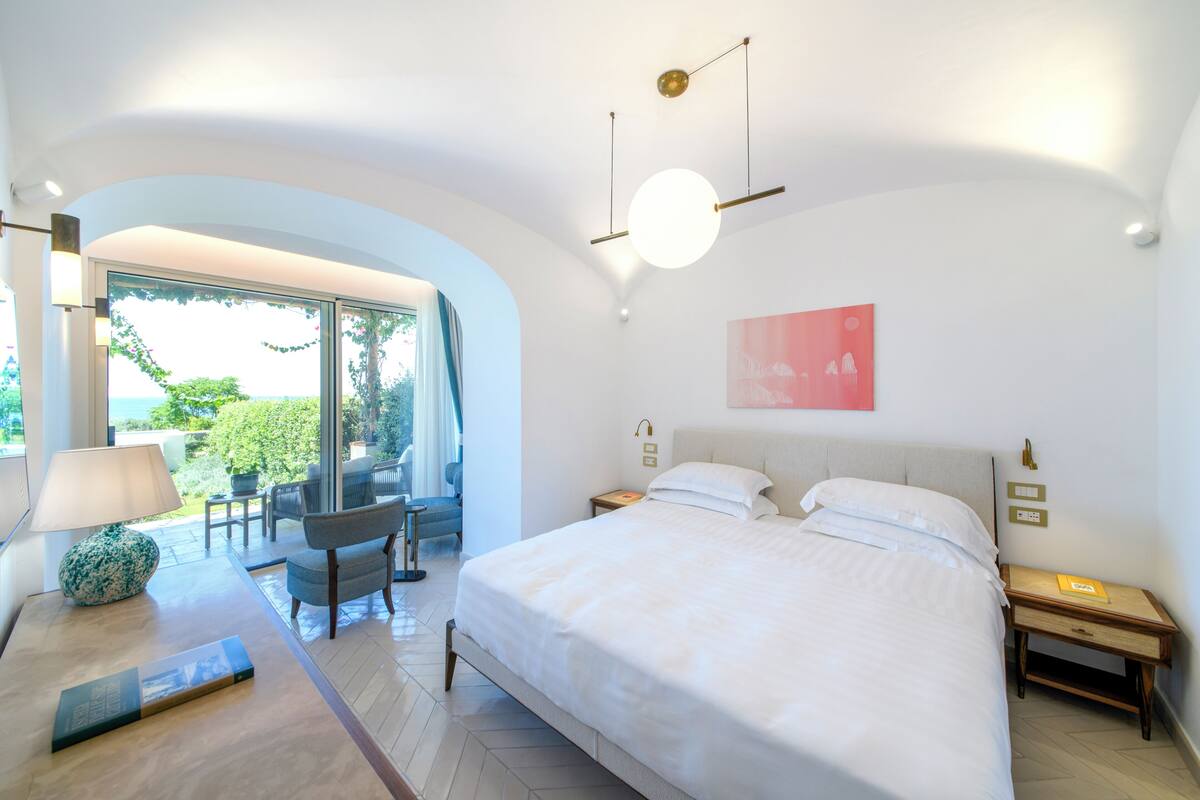 A modern bedroom with a large bed, a desk with a lamp, and a view of the outdoors.