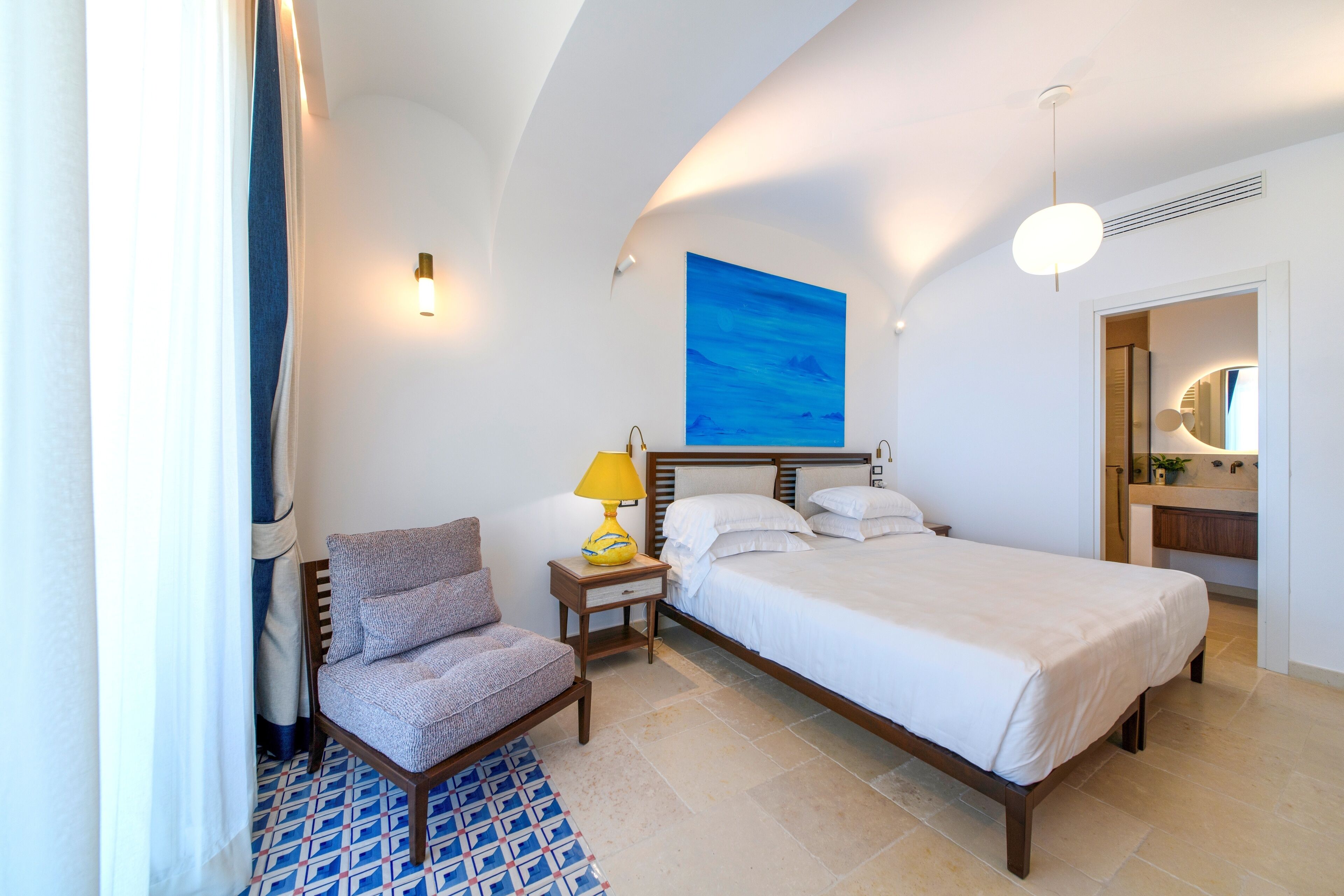 A bedroom with a bed, a chair, a nightstand with a lamp, a painting on the wall, and a bathroom visible through an open door.