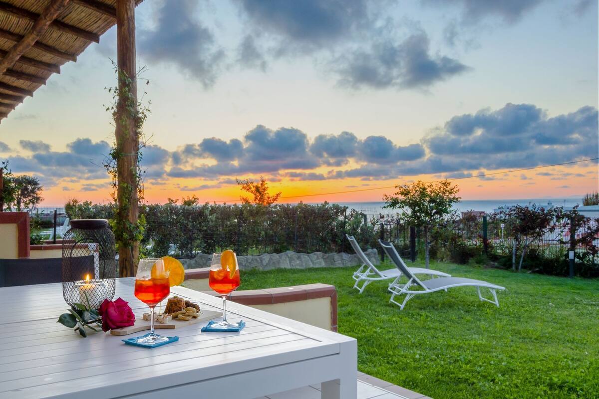 A table set for two with drinks and a candle, overlooking a grassy area and a sunset.