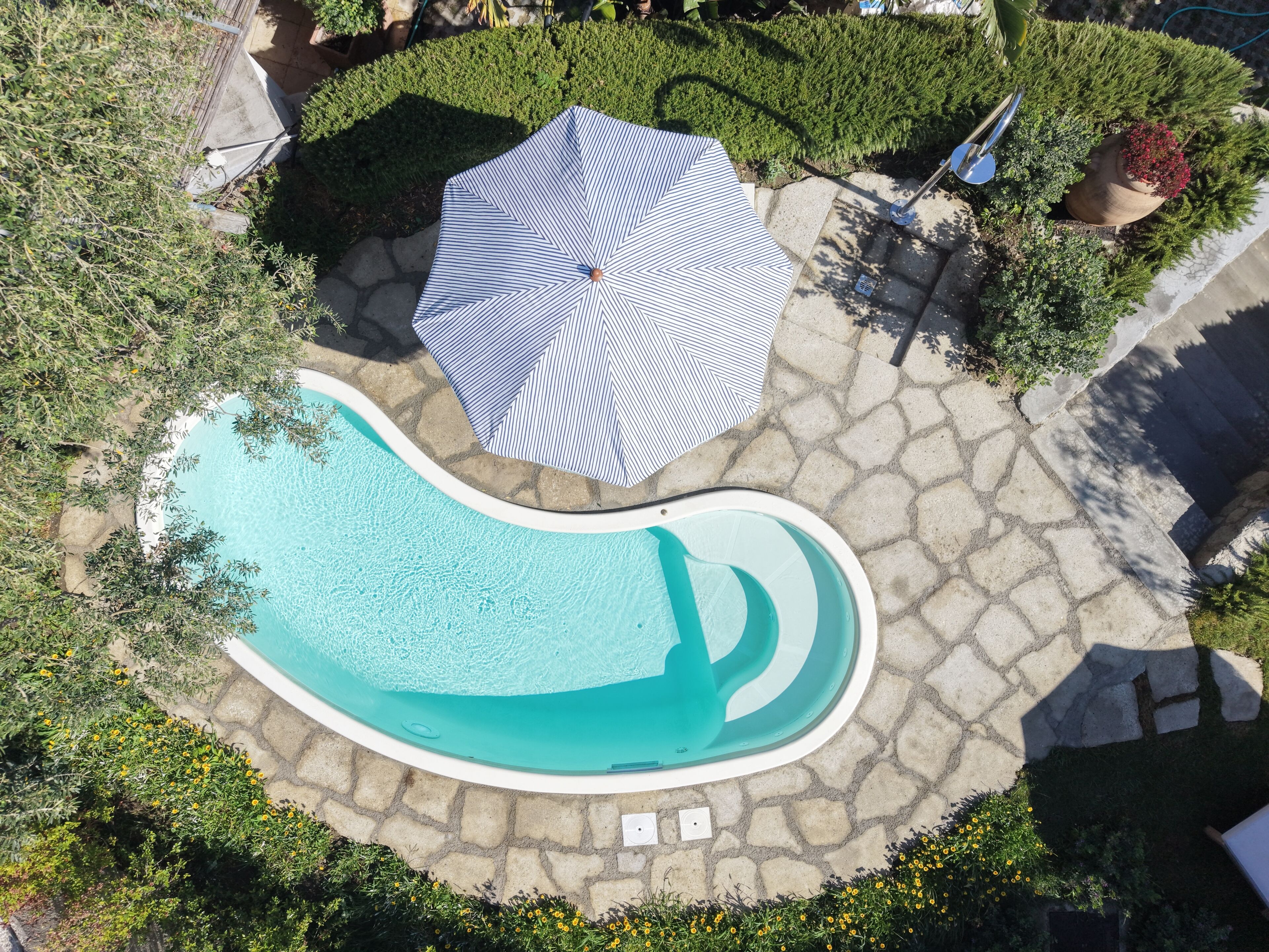 An aerial view of a swimming pool with a stone deck, surrounded by greenery and a striped umbrella.