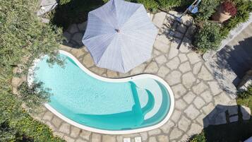 An aerial view of a swimming pool with a stone deck, surrounded by greenery and a striped umbrella.