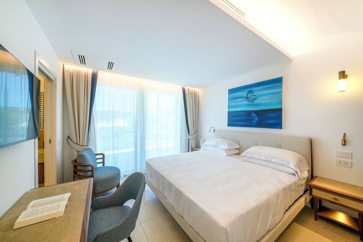 A hotel room with a bed, a desk, a chair, a TV, and a painting on the wall.