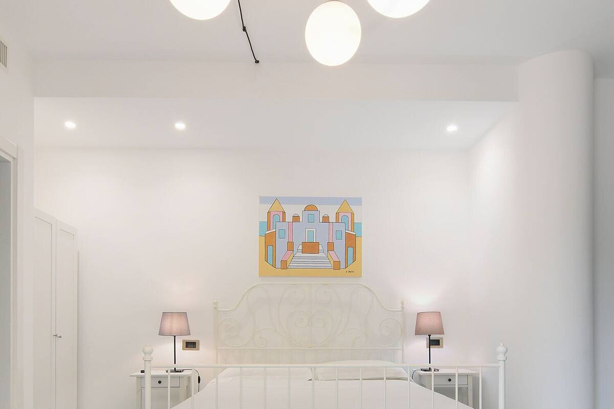 A bedroom with a white bed, two bedside lamps, a wall-mounted painting, and a tiled floor.