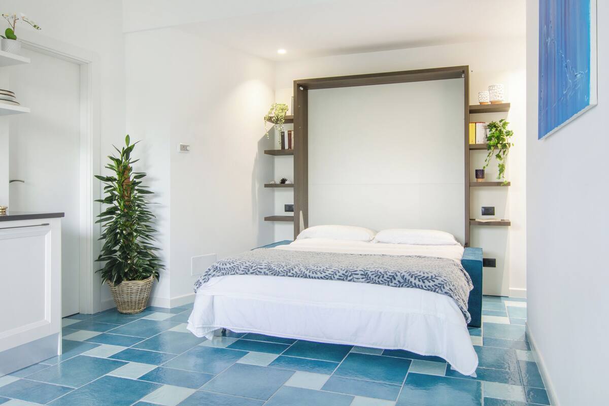 A bedroom with a bed, a nightstand, a bedside table, a wall-mounted shelf, and a potted plant.