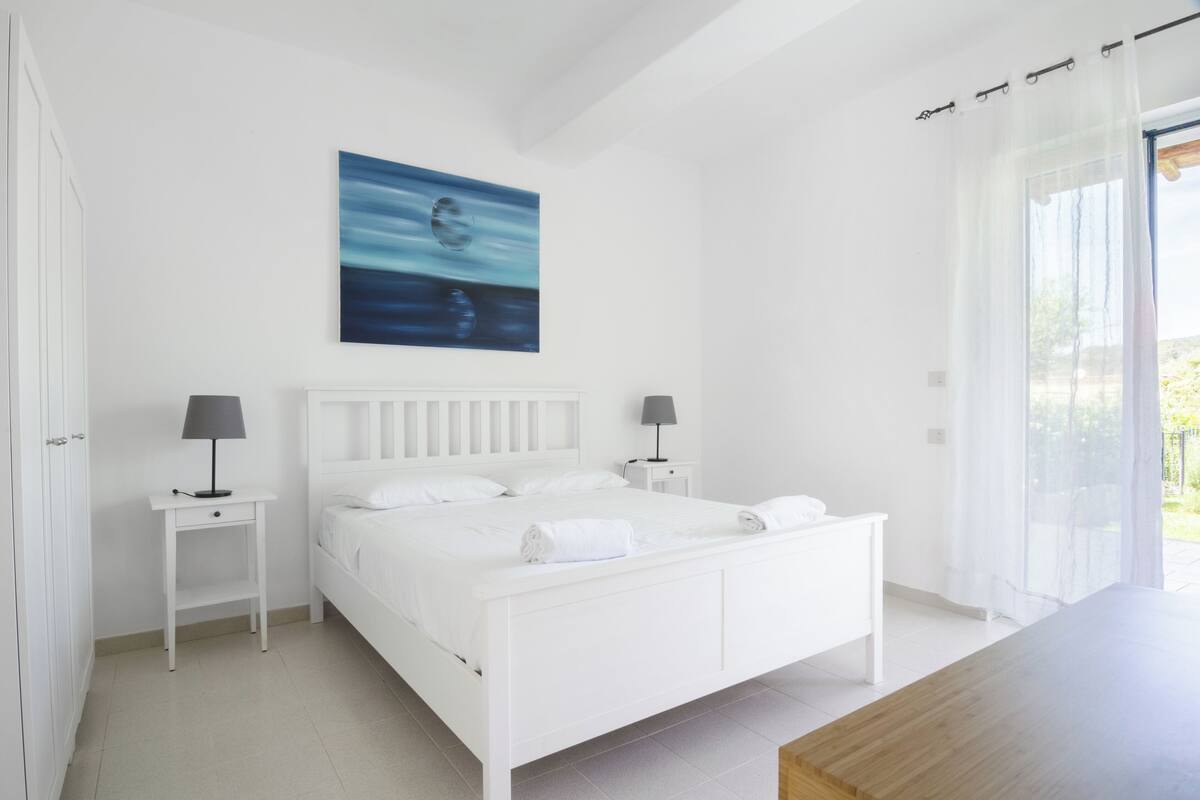 A white bedroom with a bed, two bedside tables with lamps, a painting on the wall, and a view through a window.
