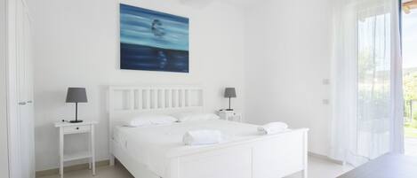 Un dormitorio blanco con una cama, dos mesitas de noche con lámparas, un cuadro en la pared y vistas a través de una ventana.