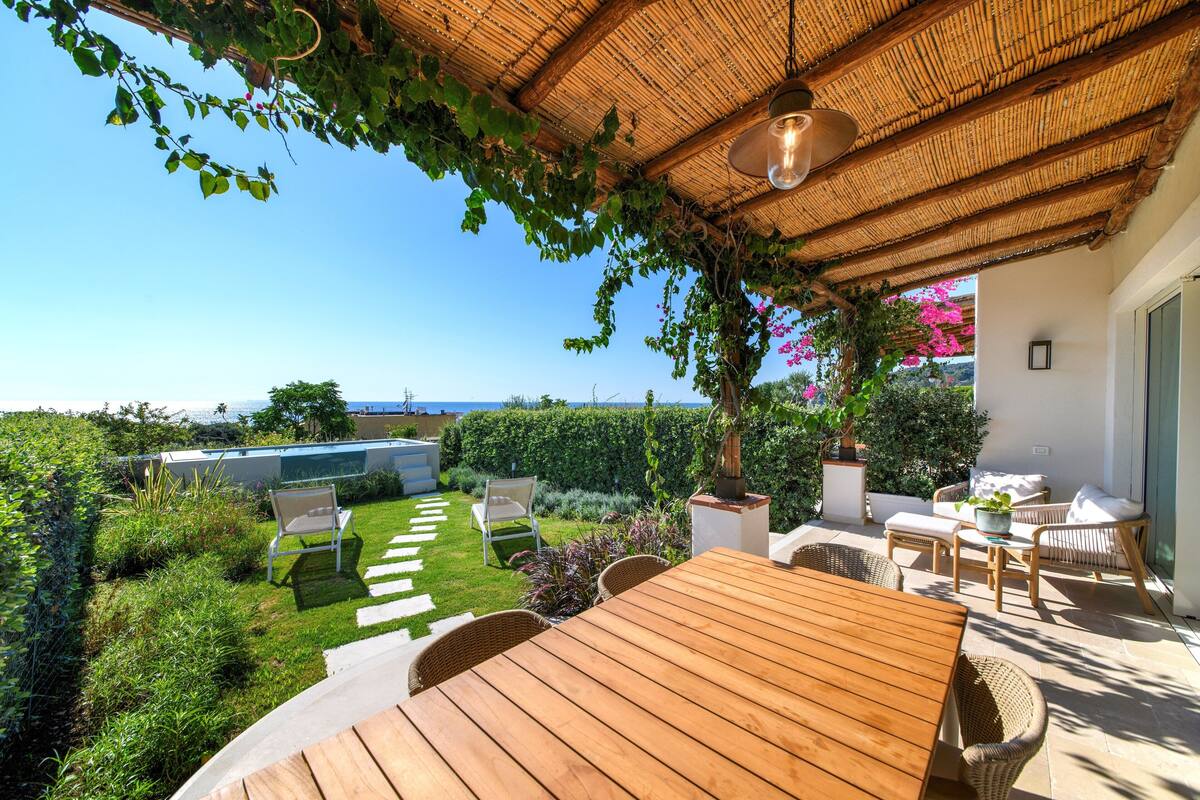 A wooden table and chairs under a thatched roof, with a view of a pool and garden.
