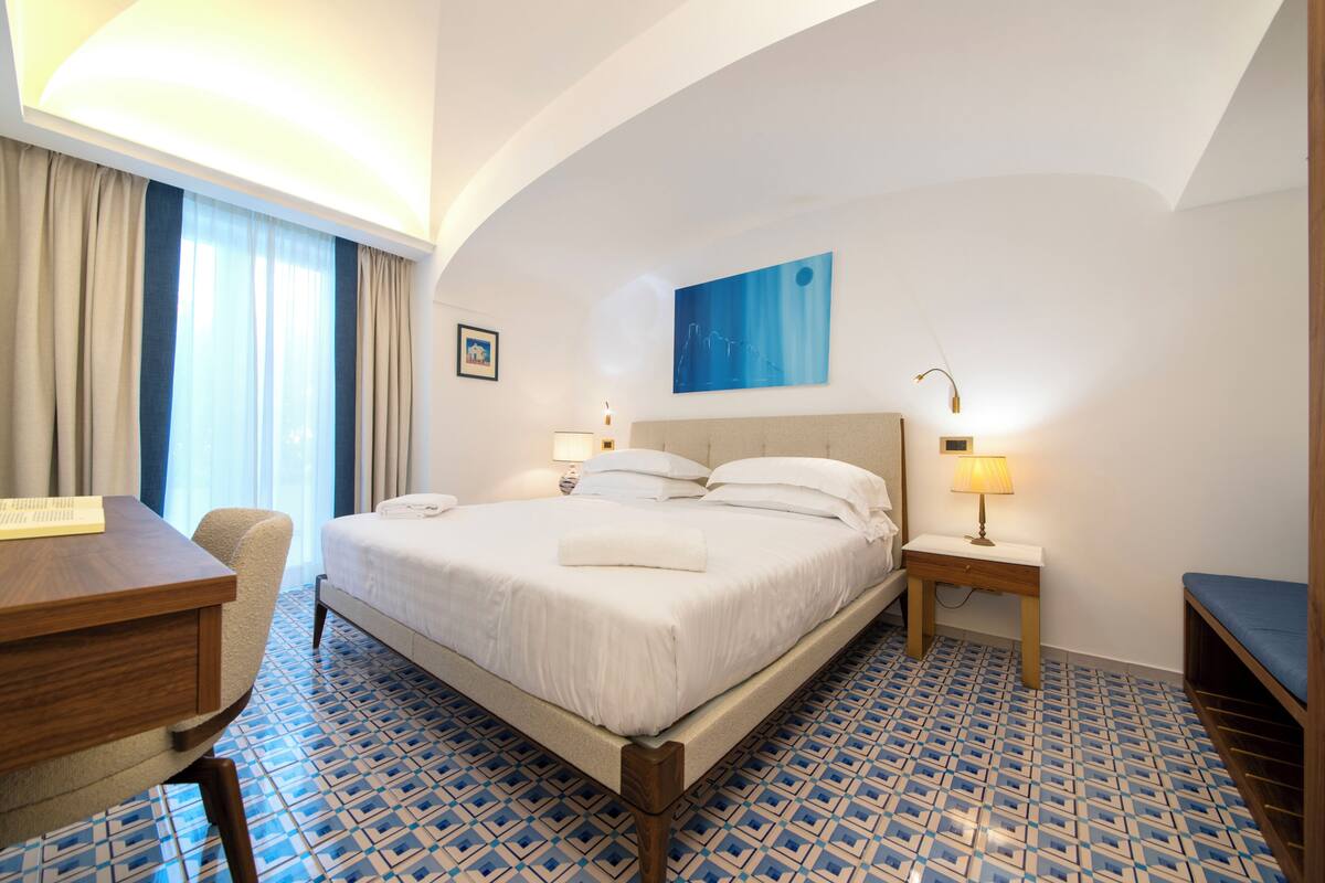A hotel room with a bed, a desk, a chair, a nightstand, and a lamp.