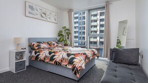 Et moderne soveværelse med en stor seng, en grå sofa, et sengebord med lampe, en potteplante og et vindue med udsigt til en bygning.