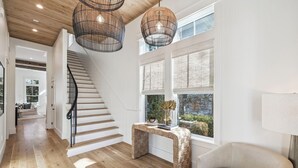Una scala conduce a una piattaforma con un tavolino consolle intrecciato e una sedia, un'ampia finestra con vista su del verde, e un lampadario.