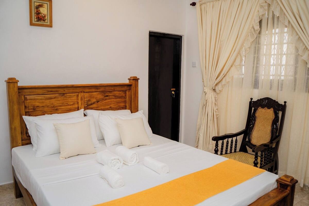 A neatly made bed with white linens and a yellow bedspread, a wooden headboard, a dark wooden chair with a woven seat, and a door with a black frame.