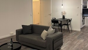 Una sala moderna con un sofá gris, una mesita redonda y un pequeño otoman. Hay un comedor con mesa y sillas, y una cocina al fondo.