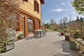 Ein zweigeschossiges Gebäude mit einer Terrasse, die einen Tisch und Stühle bietet, umgeben von Blumentöpfen und einem Kiesweg.