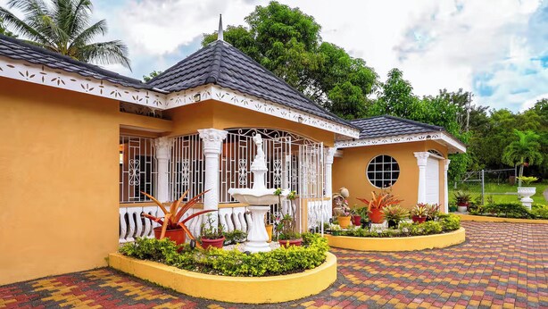 A building with a yellow facade, a tiled roof, and a paved entrance area with a fountain and potted plants. - Paradise Oasis (Tower Isle)