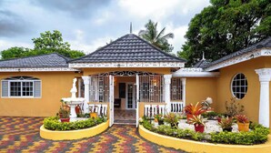 A two-story house with a tiled roof, a central entrance with a white railing, and a paved driveway with a red and yellow pattern. - Paradise Oasis (Tower Isle)