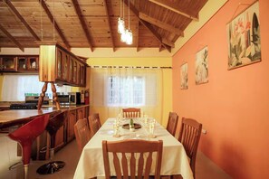 Un coin repas avec une longue table dressée pour six personnes, des chaises en bois, un luminaire suspendu et une cuisine en arrière-plan.