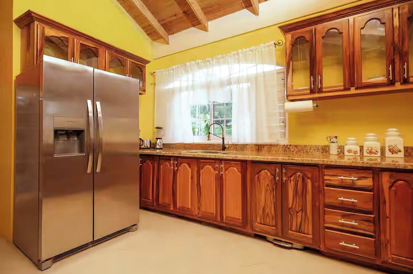 Cocina con armarios de madera, un refrigerador de acero inoxidable y una ventana con cortinas.