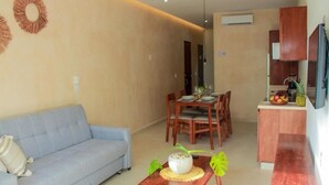 Una sala de estar con un sofá, una mesa de comedor con sillas, una pequeña zona de cocina con un refrigerador y un televisor montado en la pared.