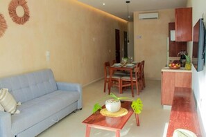 A living room with a sofa, a dining table with chairs, a small kitchen area with a refrigerator, and a wall-mounted TV. - Gama 09 Apartments - Apartment - Ground Floor (TULUM)