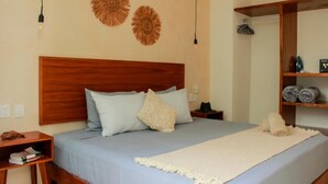 Un dormitorio ordenado con cabecera de madera, cama con cubrecama azul y manta blanca, mesita de noche con lámpara y estante con toallas enrolladas.