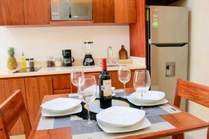 A dining table set with white plates, wine glasses, and a bottle of wine in a kitchen with wooden cabinets and a refrigerator. - Gama 09 Apartments - Apartment - Ground Floor (TULUM)