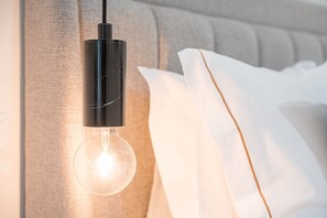 A clear light bulb hanging from a black fixture, illuminating a room with a grey upholstered headboard and white bedding. - GuestReady The 7 Seas - 'Bering Sea' Groundfloor (Porto)
