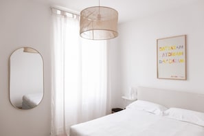 Ein schlichter Schlafzimmerraum mit Bett, einer Pendelleuchte, einem Spiegel und einem gerahmten Kunstwerk an der Wand.
