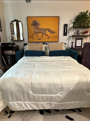 A bedroom with a large bed, a mounted TV, a framed artwork of a horse, a plant on a shelf, and a desk with a computer.
