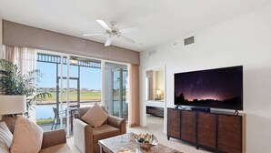 Een moderne woonkamer met een grote flatscreen televisie, een plafondventilator en uitzicht op een golfbaan door grote ramen.