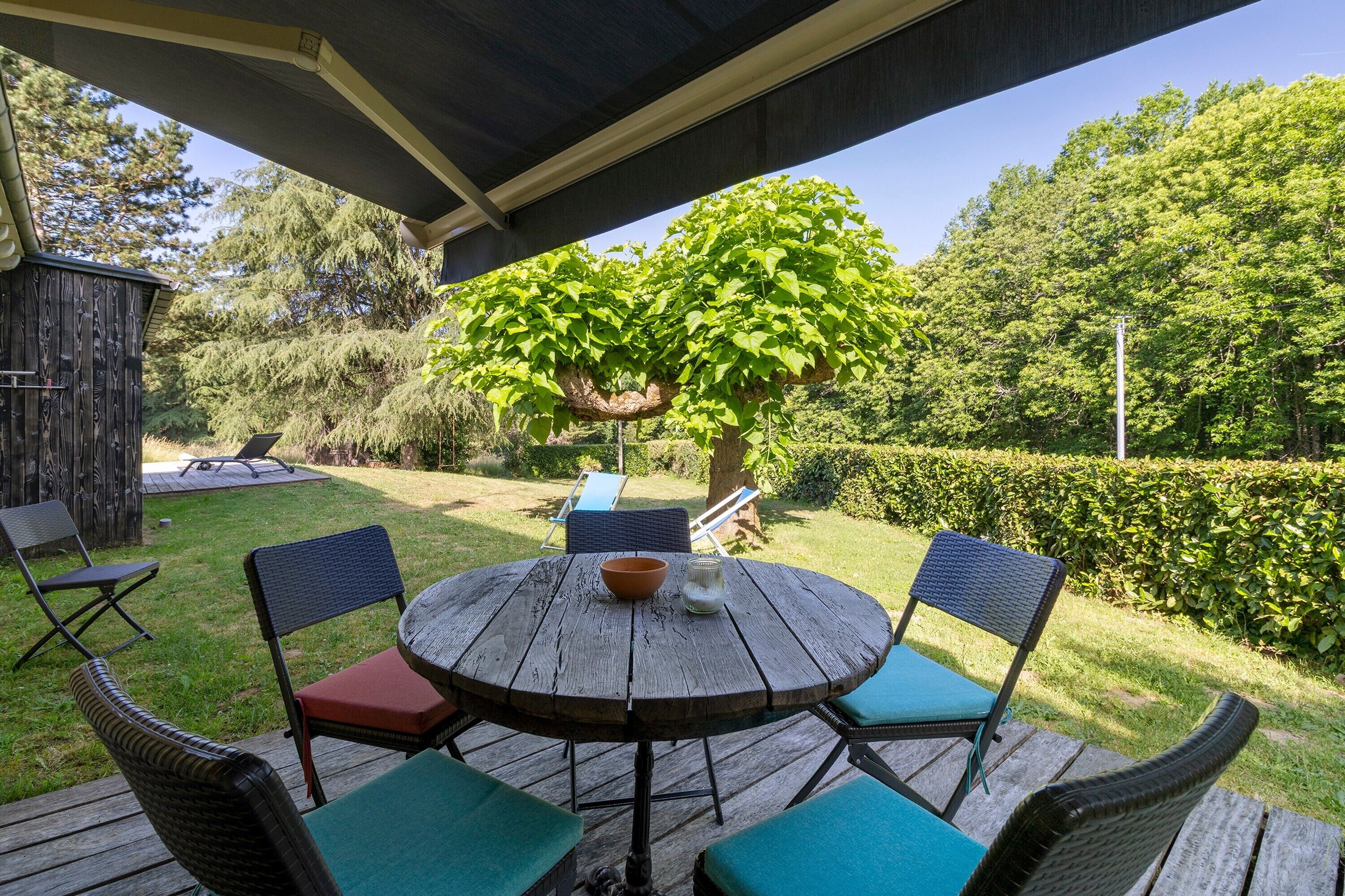 Una terraza de madera con una mesa redonda y sillas, rodeada de follaje y un árbol.