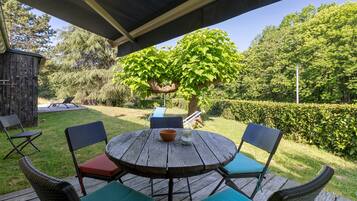 A wooden deck with a round table and chairs, surrounded by greenery and a tree.