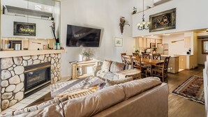 A spacious living room with a stone fireplace, a large flat-screen TV, a dining area with wooden furniture, and a kitchen in the background. - New Listing! Ski In/Walk to the Lift (Breckenridge)