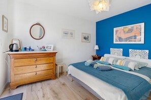 Un dormitorio con cómoda de madera, cama con ropa de cama azul, pared de acento azul y una obra enmarcada sobre la cama.
