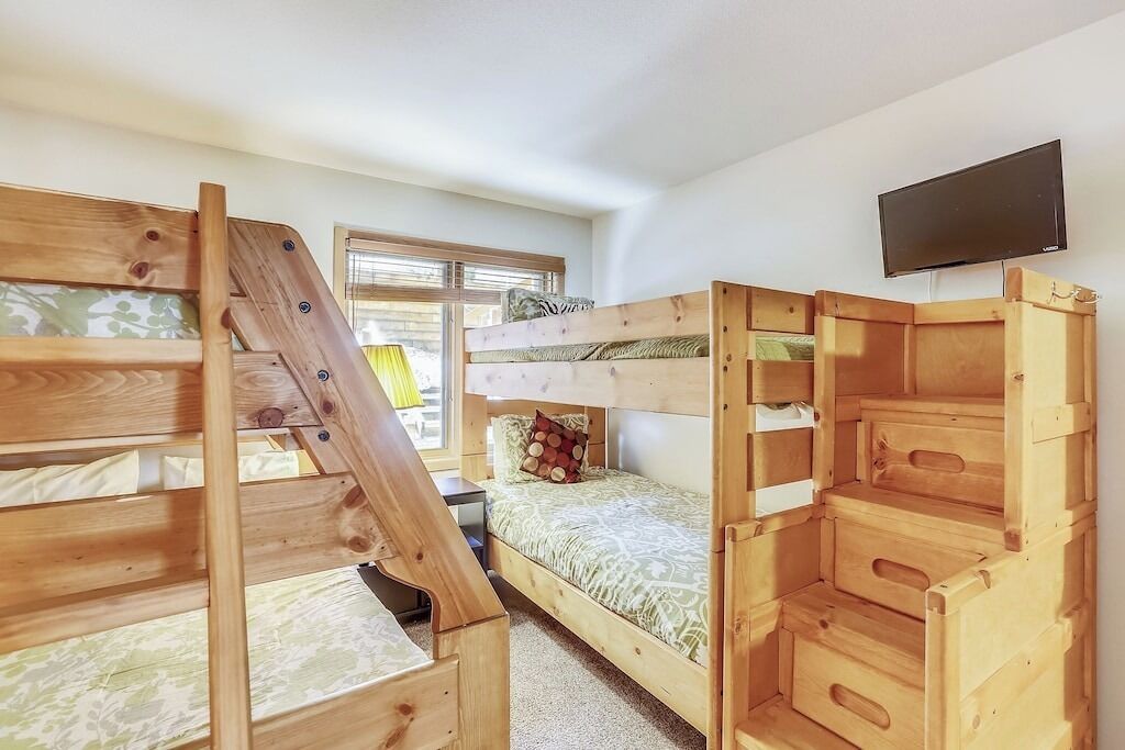A room with bunk beds, a television mounted on the wall, and a window with blinds.