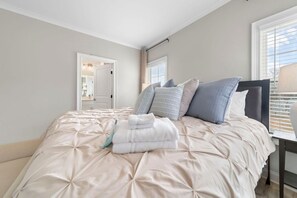 Ein ordentlich bezogenes Bett mit beiger Decke und Kissen, ein Nachttisch mit Lampe und ein Fenster mit Jalousien.