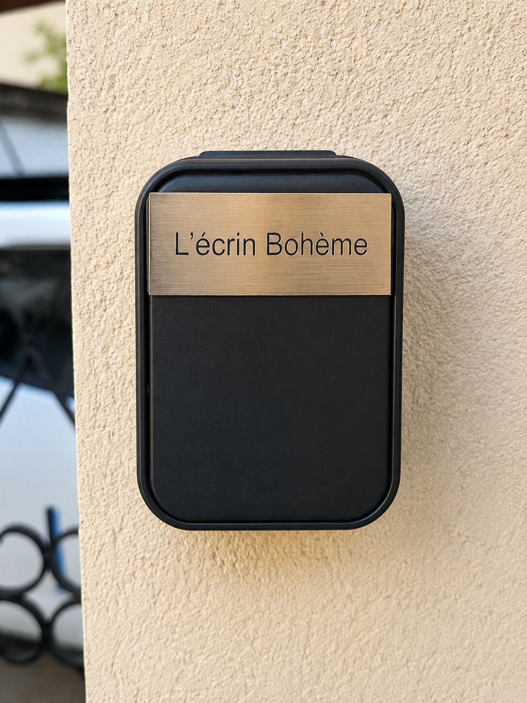 Een zwart, rechthoekig object met een messing plaquette waarop 'L'écrin Bohème' staat, bevestigd aan een beige muur met een structuur.