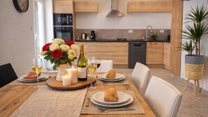 Une table à manger dressée avec des assiettes, des verres à vin, une bougie et un bouquet de fleurs, dans une cuisine moderne aux meubles en bois et agrémentée d’une plante en pot.
