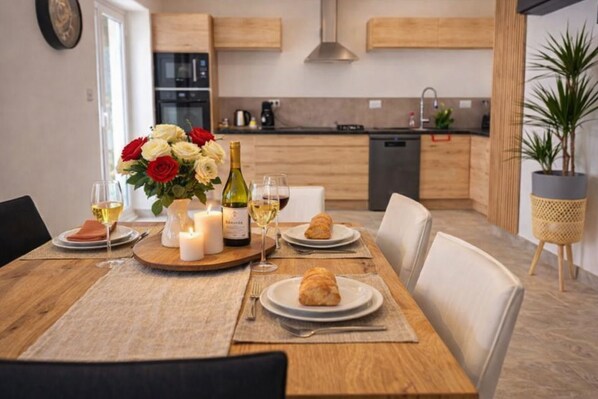 Une table à manger dressée avec des assiettes, des verres à vin, une bougie et un bouquet de fleurs, dans une cuisine moderne aux meubles en bois et agrémentée d’une plante en pot.