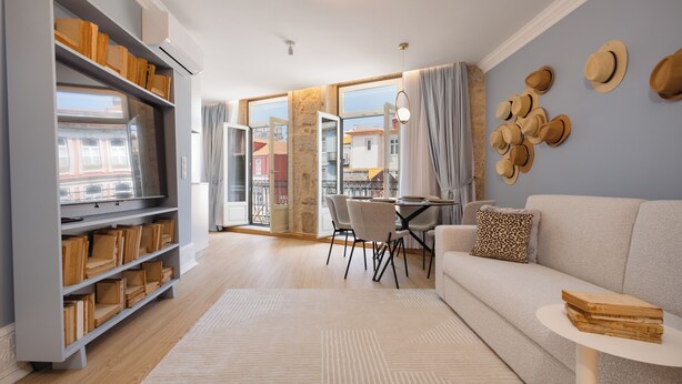 A modern living room with a sofa, dining table, and chairs, a bookshelf, and a view through large windows.