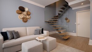 A modern living room with a sofa, ottoman, and a round coffee table. A staircase with glass railings leads to a door.