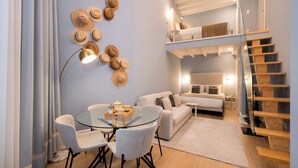 A modern living room with a glass table, grey chairs, a sofa, a staircase, and a wall-mounted hat display.